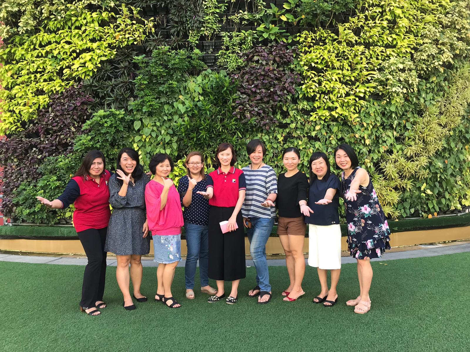 Nine women posing and smiling in a row on grass in front of a large green living plant wall.